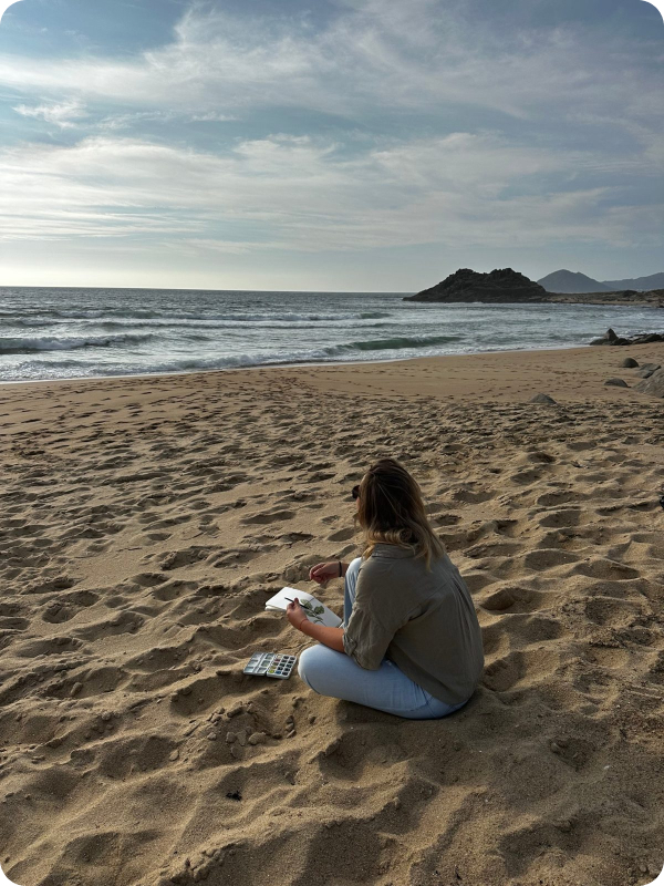 Caro sitting on a beach, drawing in a sketchbook